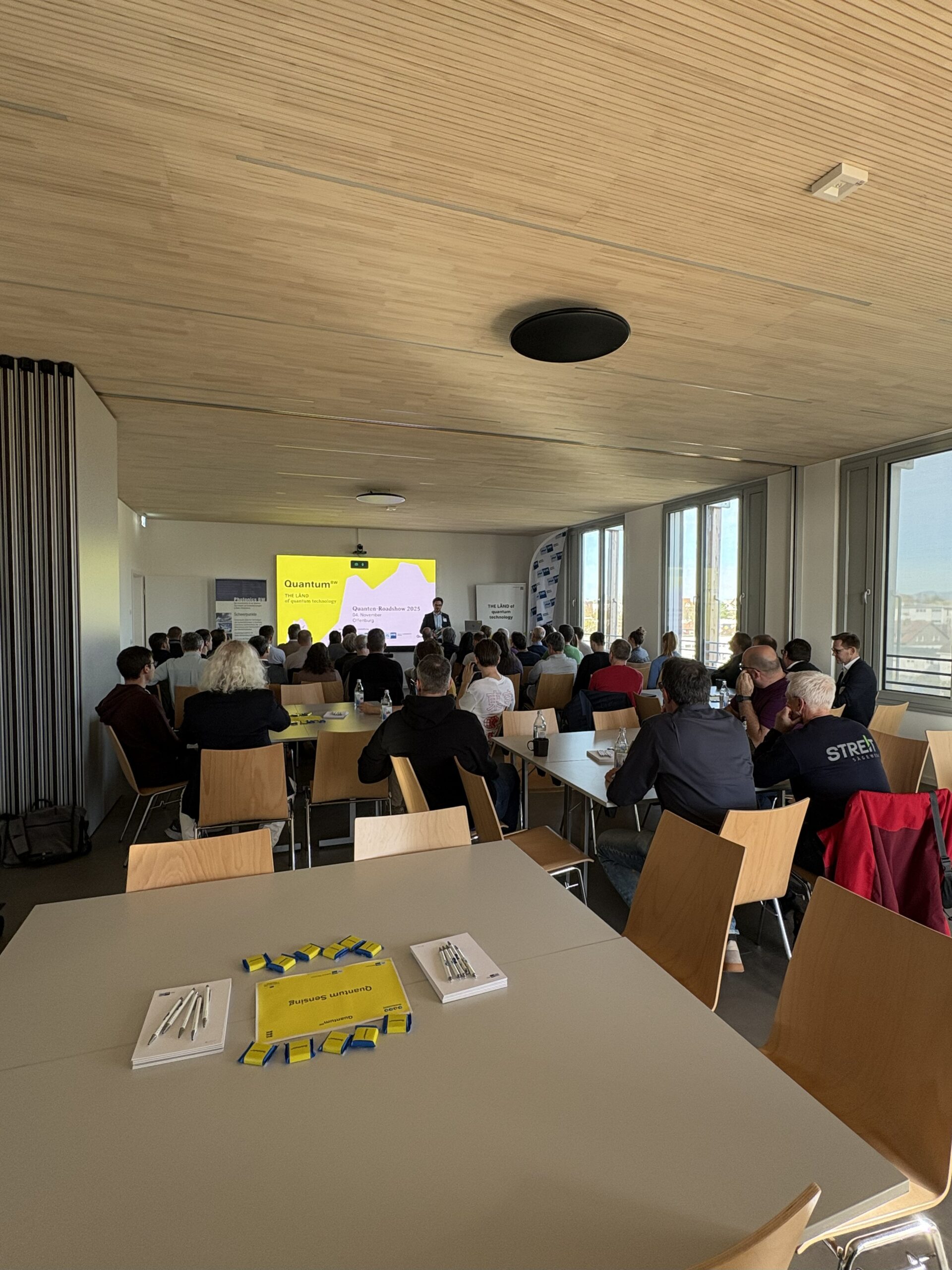 Bild einer Veranstaltung aus der Perspektive des Auditoriums mit Blick auf eine Bühnenfläche mit einer Präsentation und einem Redner.