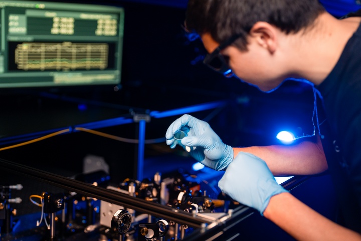 Experimenteller Aufbau eines Quantennetzwerkknotens, bei dem ein junger Forschender vor einem in blaues Licht getauchten optischen Aufbau steht und eine Linse in der Hand hält.